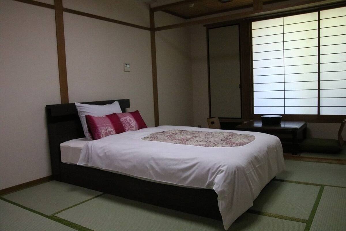 Une chambre de style japonais traditionnel, avec un sol recouvert de tatamis, un lit avec une literie blanche et des oreillers rouges, une tête de lit en bois et une fenêtre coulissante de type shoji.