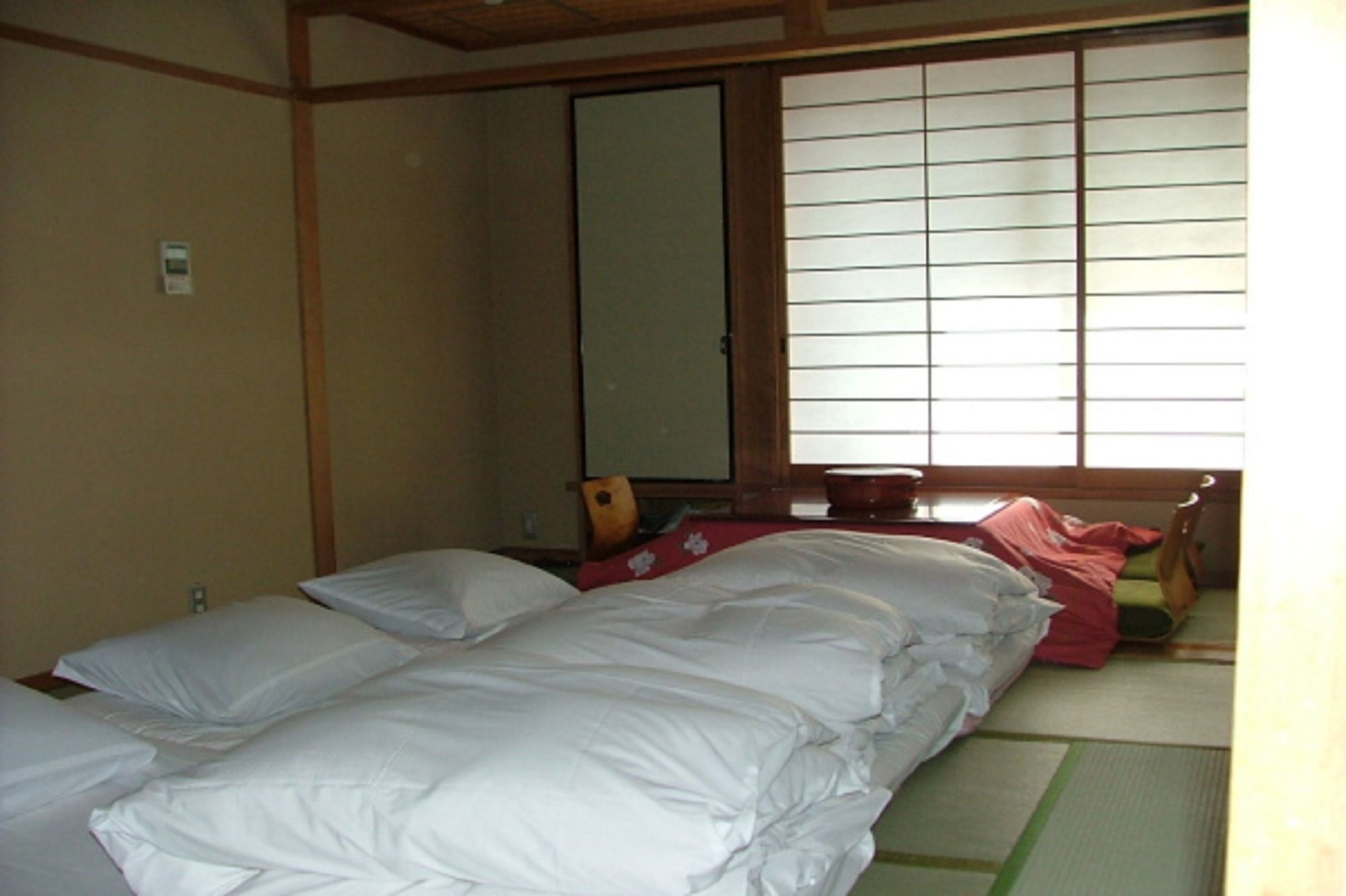 A traditional Japanese-style room with tatami flooring, a low wooden bed with white bedding, a sliding shoji screen window, and a small table with a red cloth.