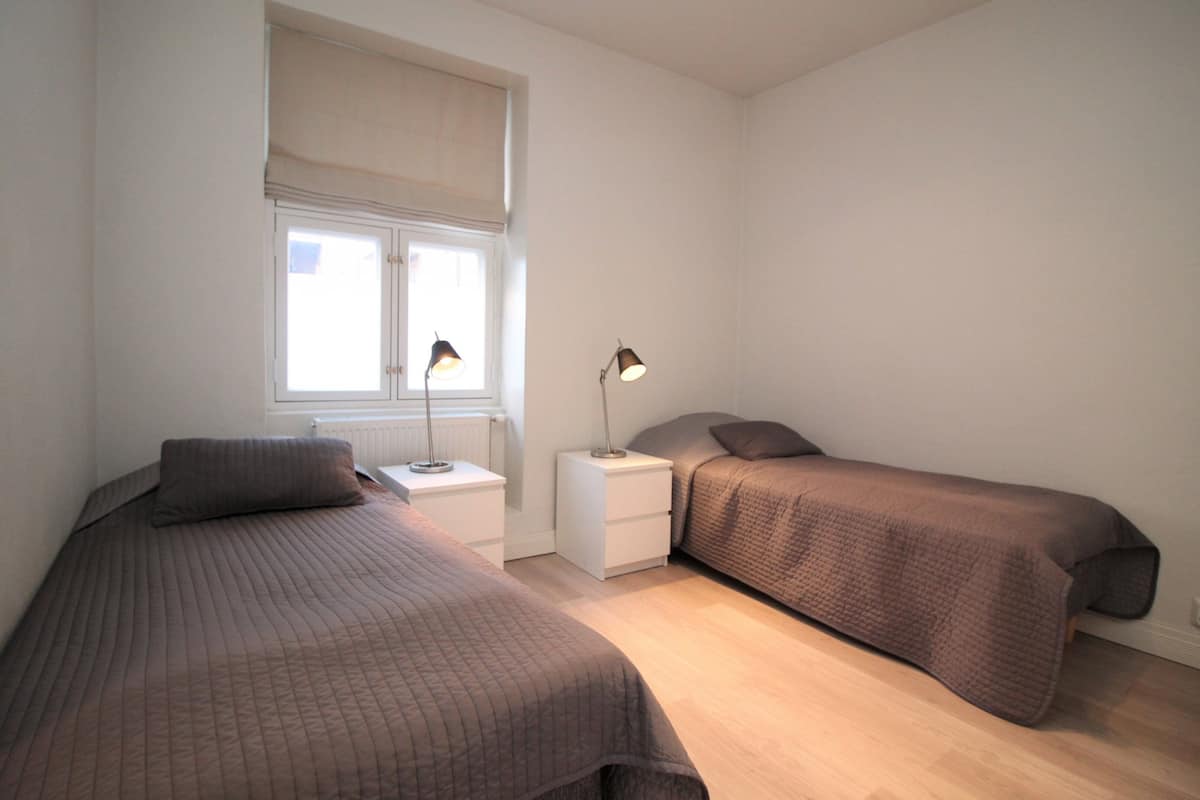 A bedroom with two beds, a window with blinds, bedside lamps, and a radiator.