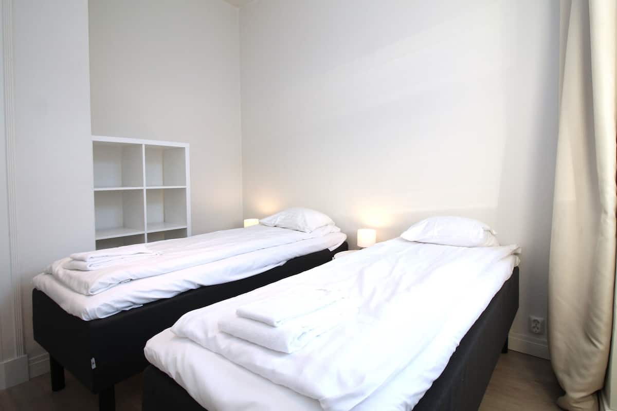 Two single beds with white linens, a white shelving unit, and bedside lamps in a room with white walls.
