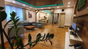 A modern living room with a sofa, armchairs, a coffee table, and a TV. There is a dining area in the background and a kitchen visible through an open door. - Hotel Crocus (Štrba)