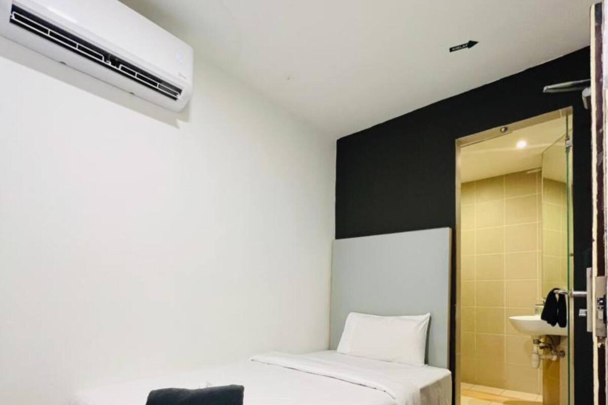 A single bed with white bedding, a black headboard, and a black pillow. There is a wooden floor and a white air conditioning unit mounted on the wall.