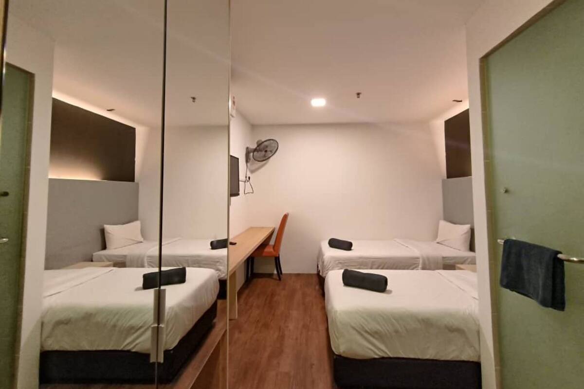A compact hotel room with two single beds, a wooden table, a chair, a wall-mounted fan, and a small window.