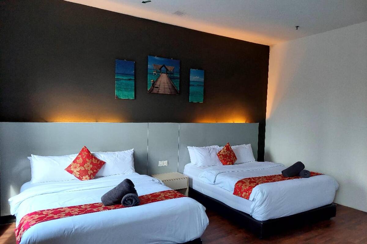 A hotel room with two beds, each with white bedding and red floral pillows. There are three framed pictures on the wall above the beds.