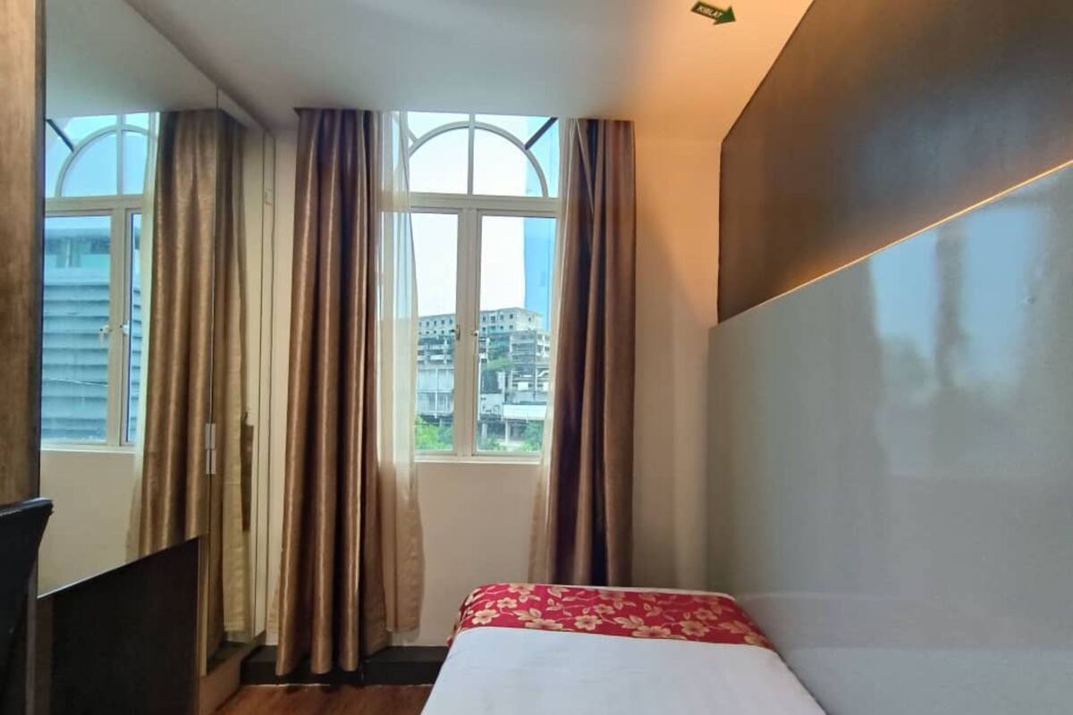 A small, neatly arranged room with a bed, a window with curtains, and a view of buildings outside.