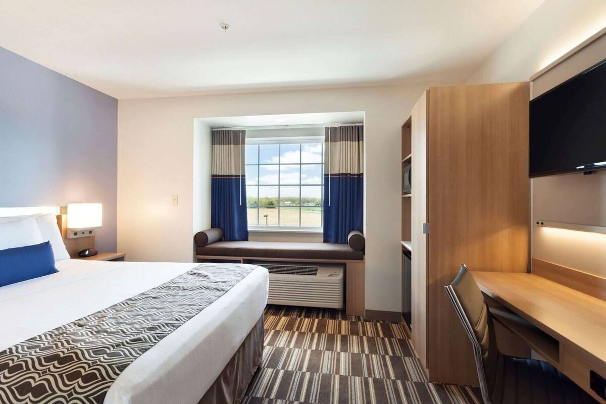 A hotel room with a bed, a desk, a TV, a window with curtains, and a view of the outdoors.