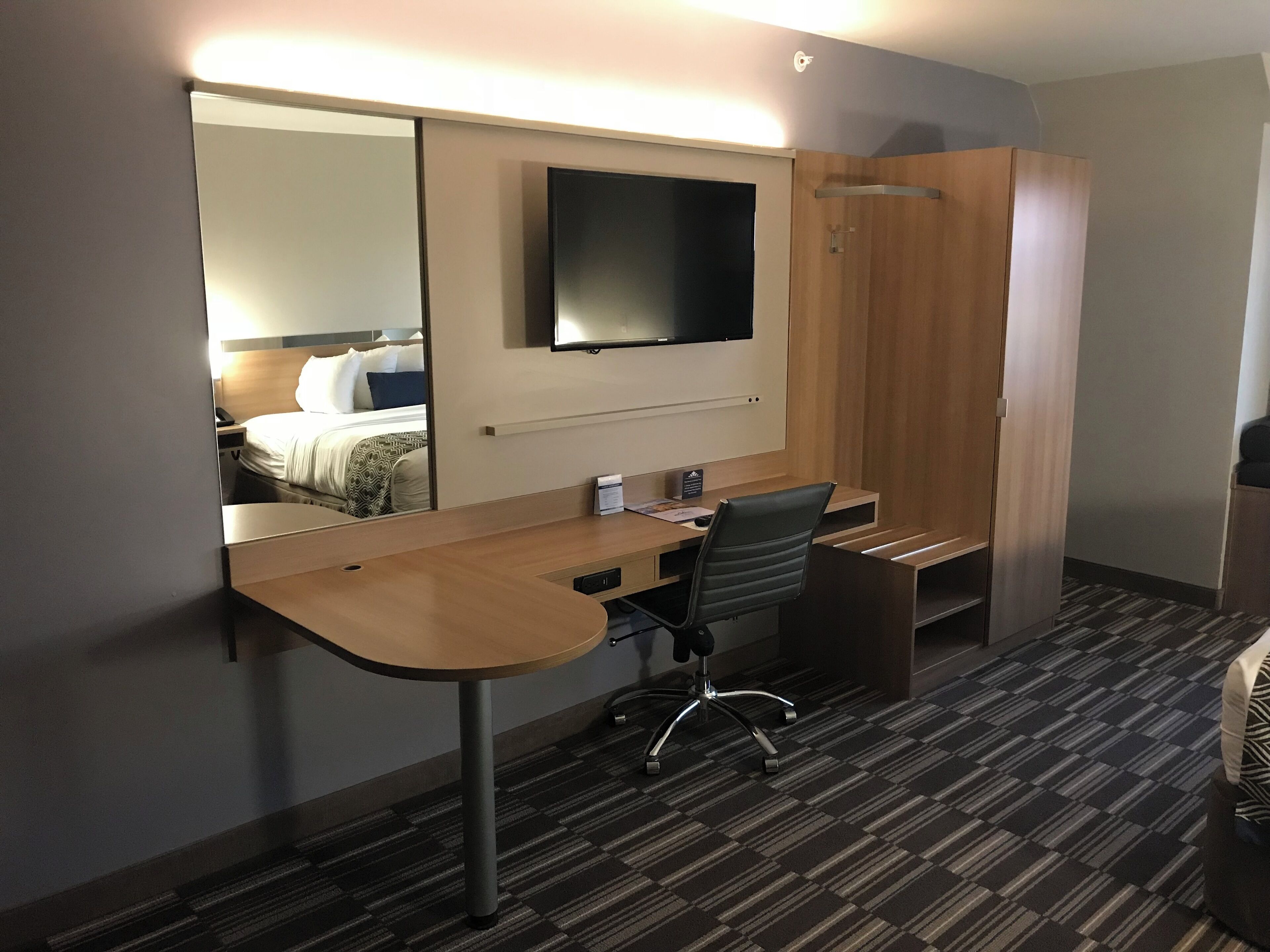 A hotel room with a desk, chair, and a large mirror reflecting a bed.