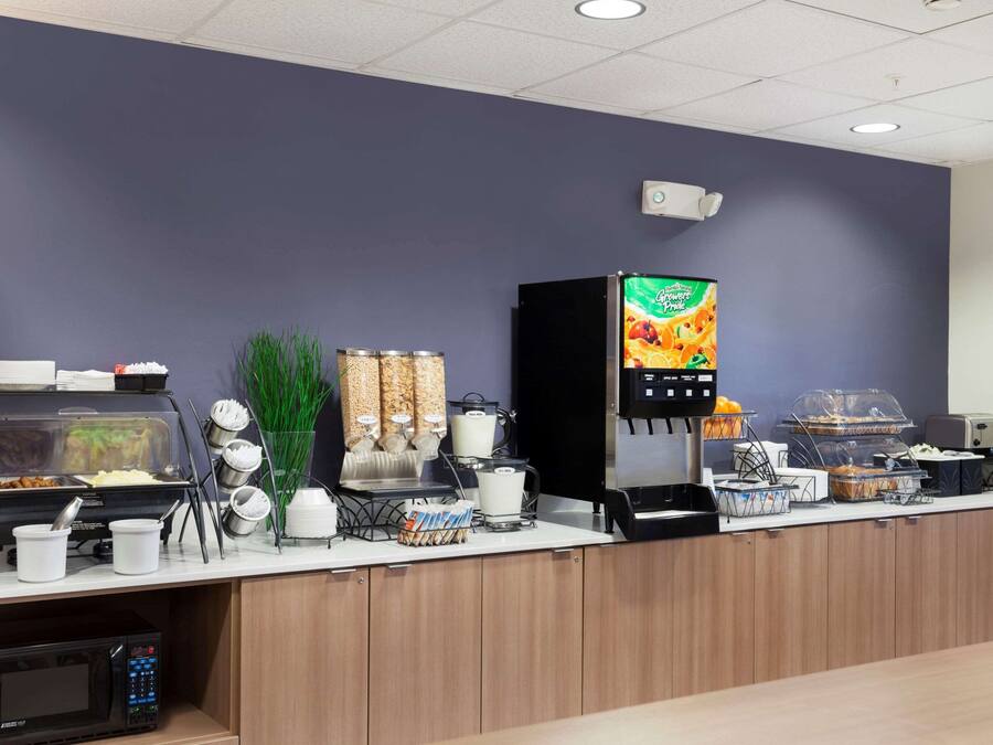 A self-service breakfast station with a coffee machine, cereal, and a juice dispenser.