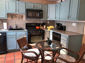 A kitchen with light blue cabinets, a black stove, a microwave, and a coffee maker. There is a wicker chair and a glass table with a vase of flowers. - Casas de Guadalupe (Santa Fe)