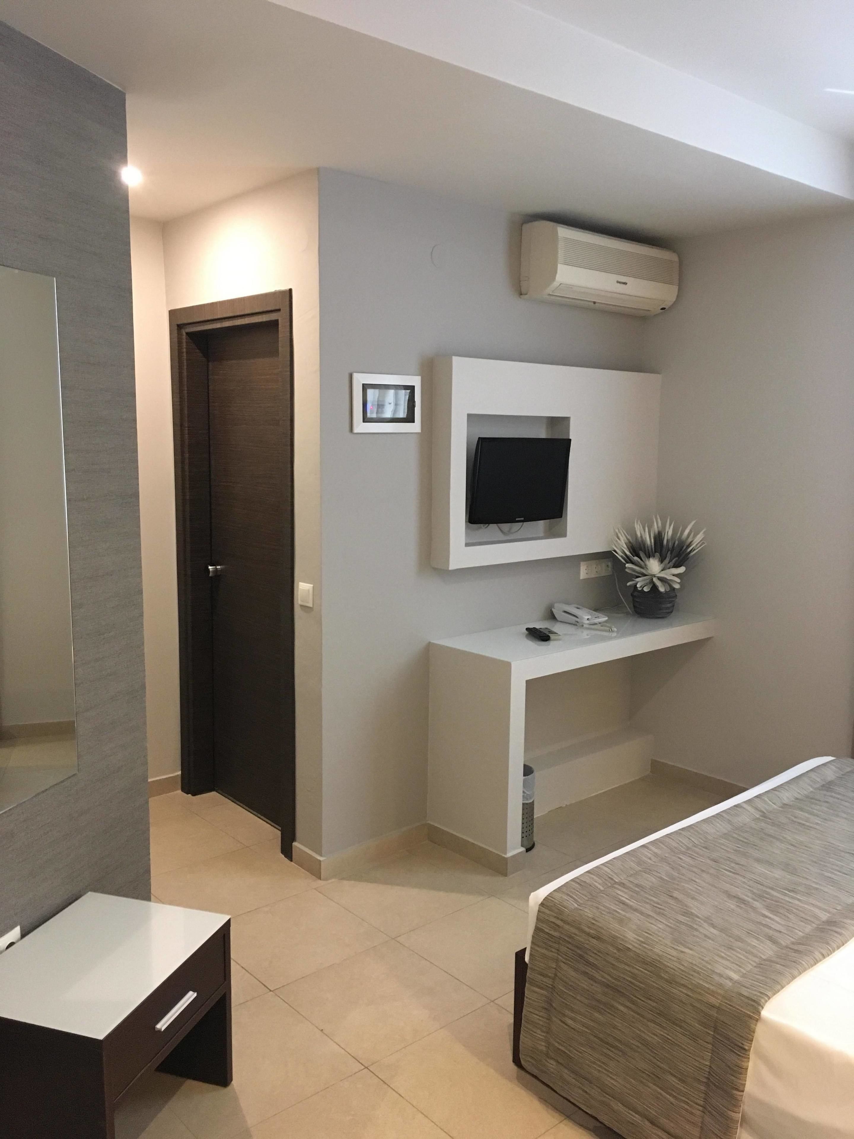 Un dormitorio moderno con una cama, un televisor montado en una consola blanca, una puerta y una pequeña planta en maceta.