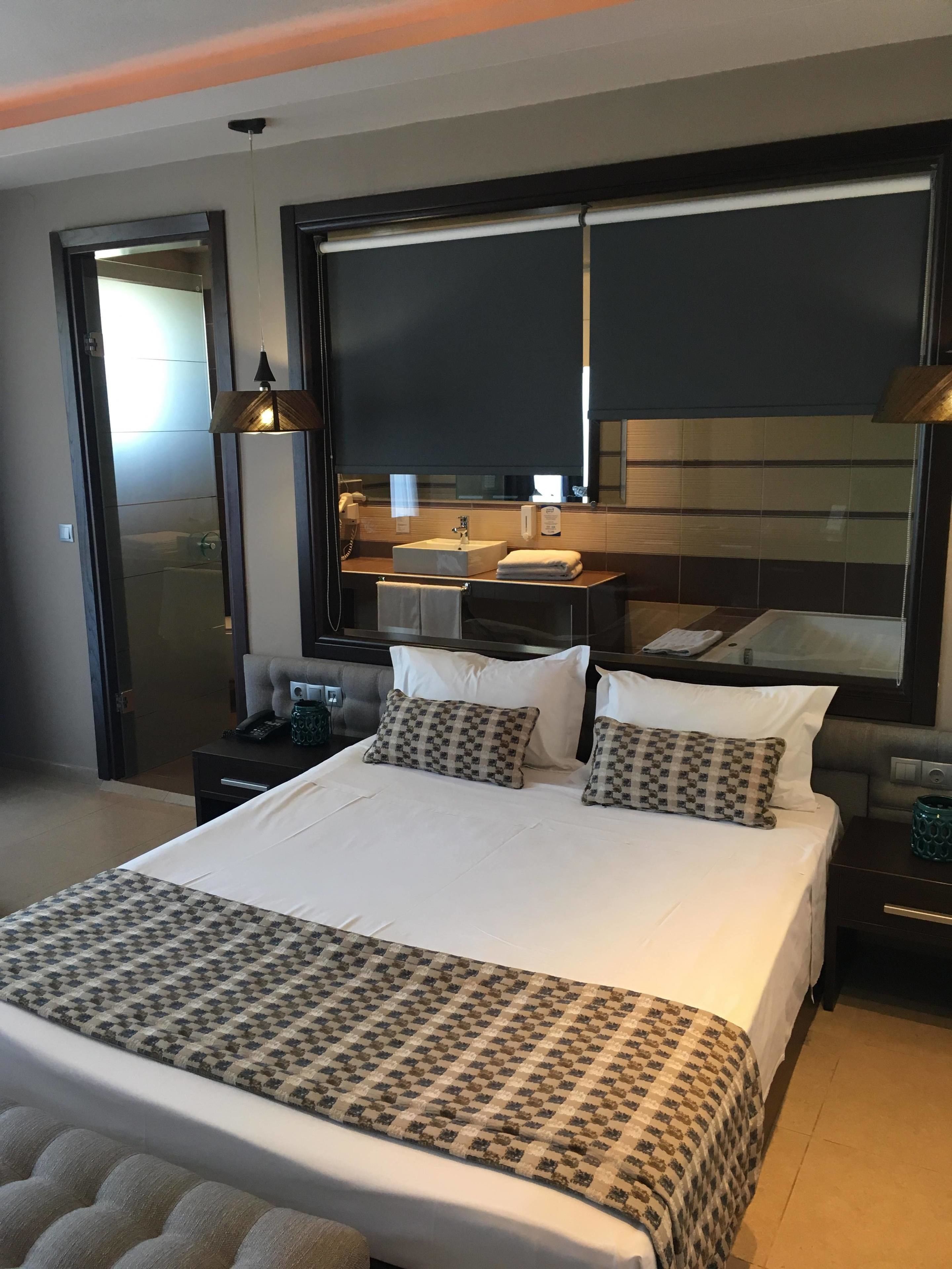 A modern hotel room with a large bed, a bedside table, a nightstand, a bathroom mirror, and a window.