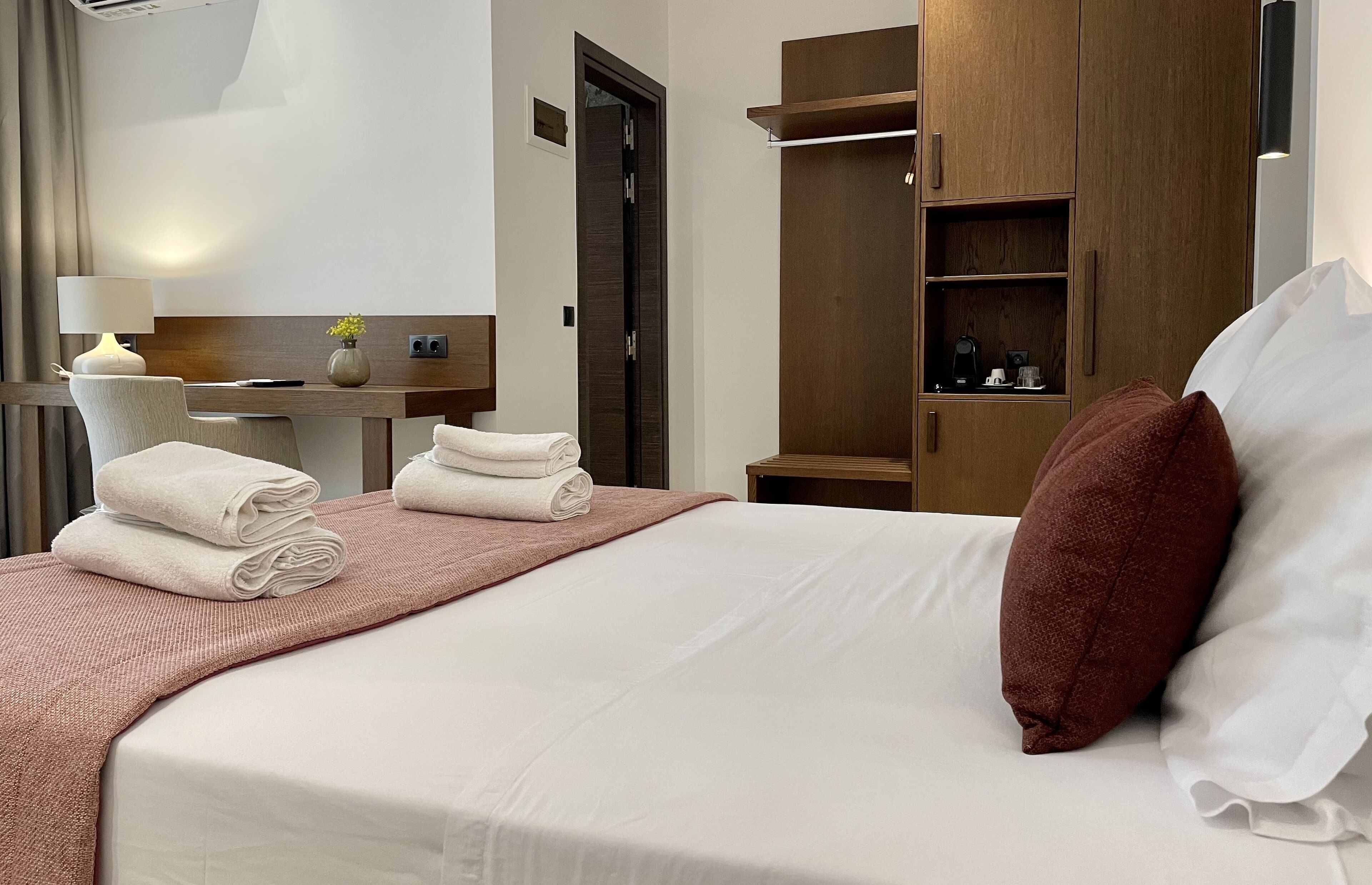 A neatly made bed with folded towels, a wooden wardrobe, a desk with a lamp, and a small plant.