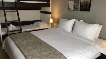 A hotel room with a bunk bed, a single bed, white linens, a bedside lamp, and framed artwork on the wall.