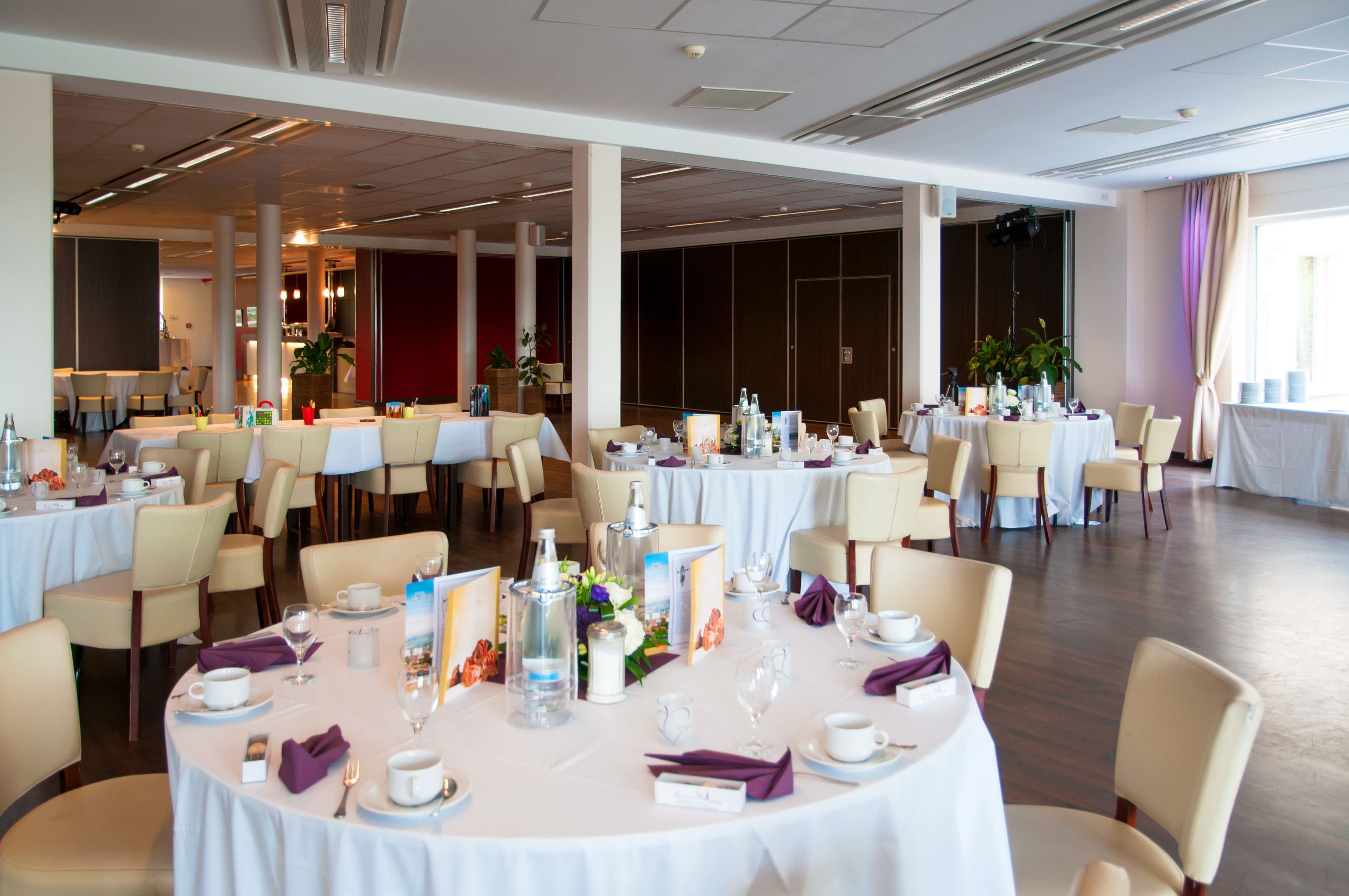 A spacious banquet hall with round tables set for dining, featuring white tablecloths and beige chairs.
