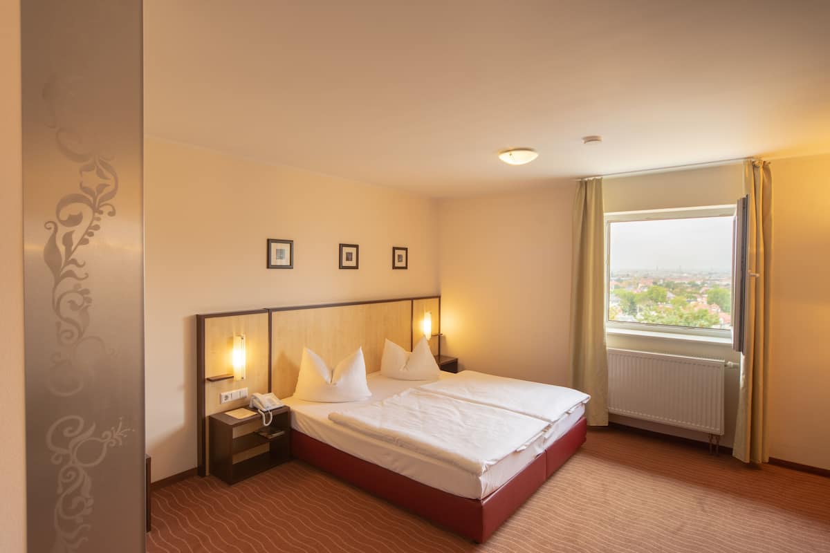 Ein Hotelzimmer mit einem großen Bett, Nachttischlampen, einem Beistelltisch, einem Fenster mit Vorhängen und Blick auf die Stadt.