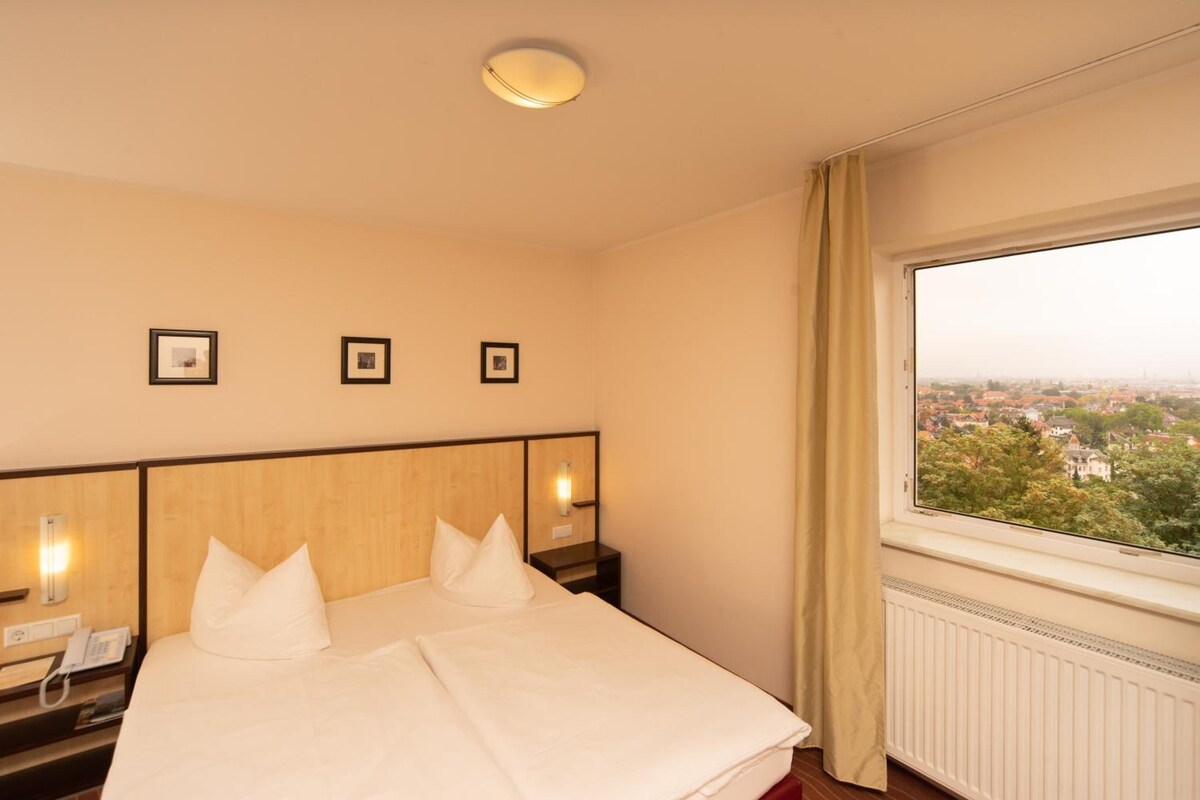 Una camera d'albergo con un letto, comodini, una finestra vista città e quadri appesi al muro.