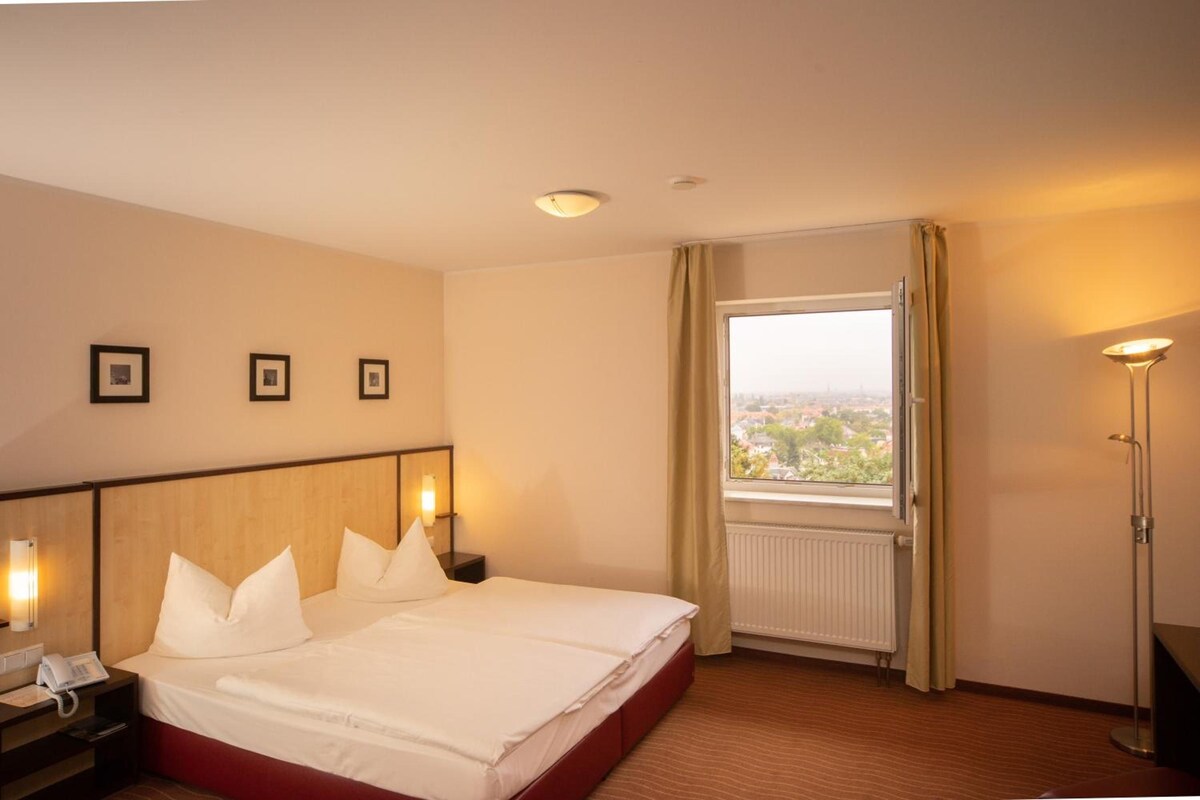 Una habitación de hotel con cama, mesitas de noche, un buró con teléfono, una ventana con cortinas y vistas de la ciudad.