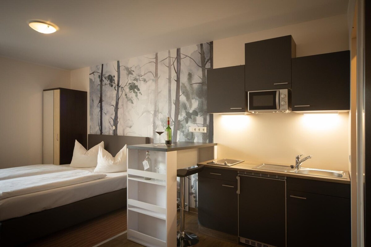Una habitación pequeña con cama, una kitchenette y televisor montado en la pared.