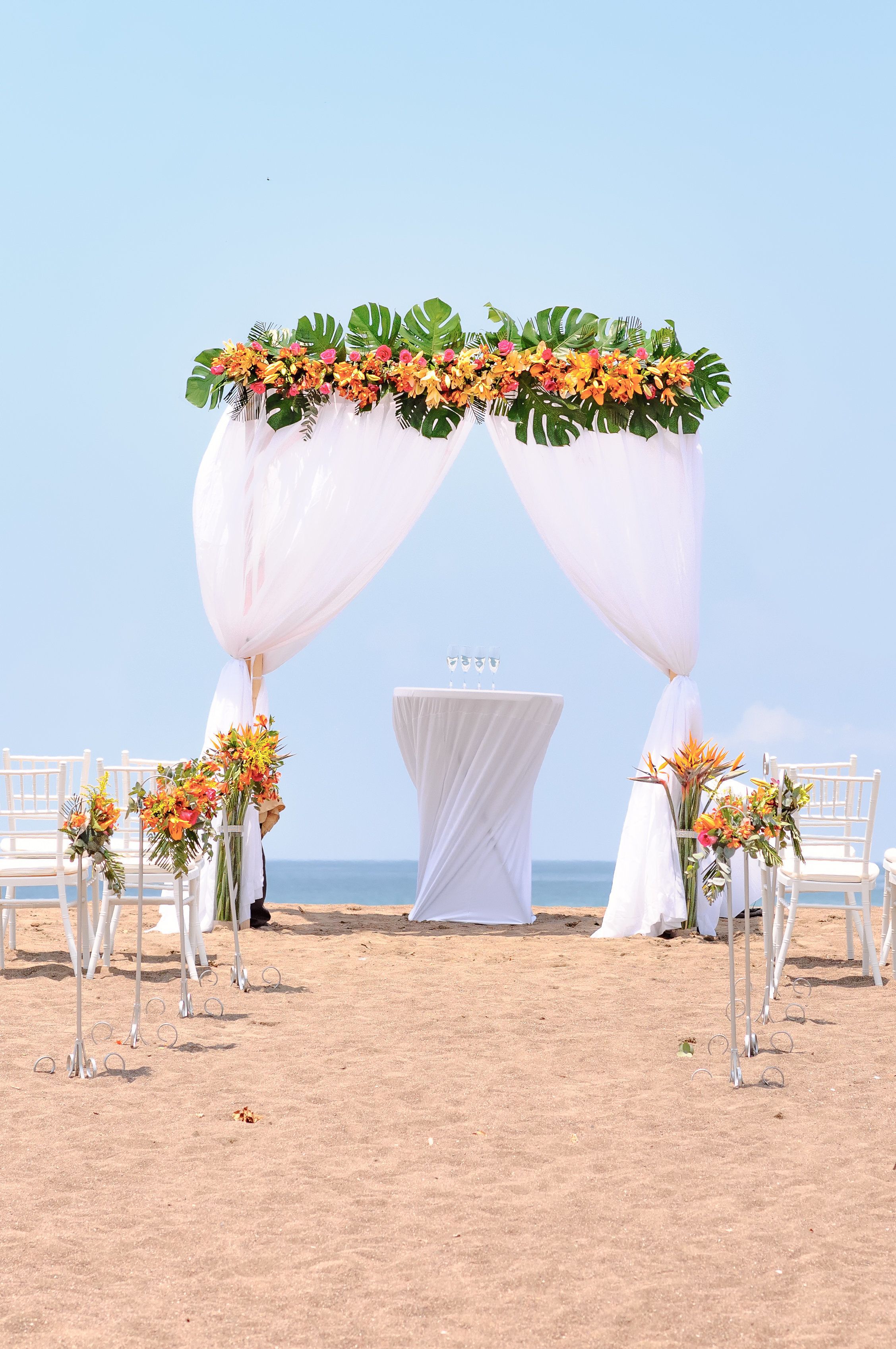 A beach wedding setup with a white arch adorned with flowers and draped fabric, white chairs, and a table with a white cloth.