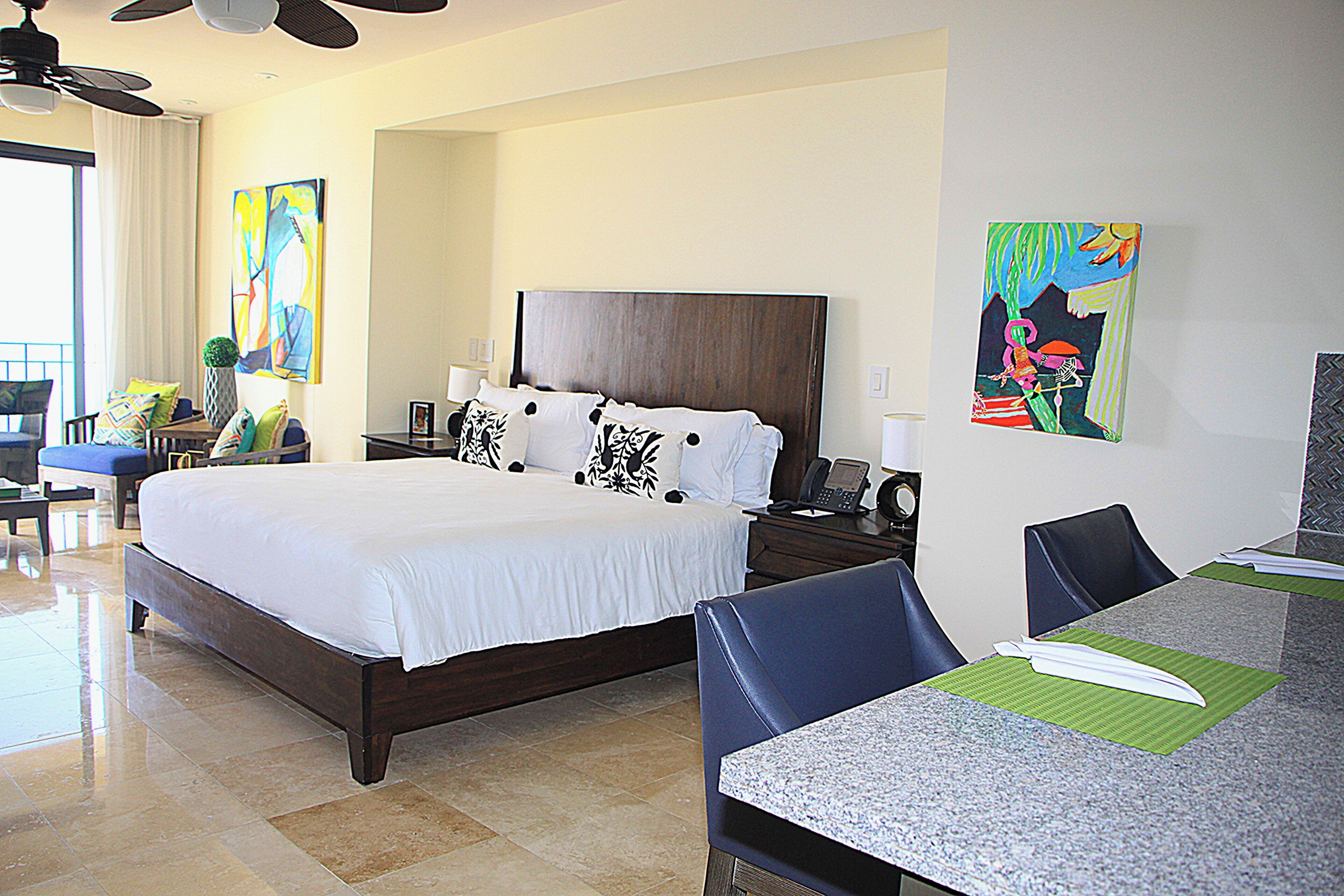 A hotel room with a large bed, a desk with chairs, and artwork on the walls.