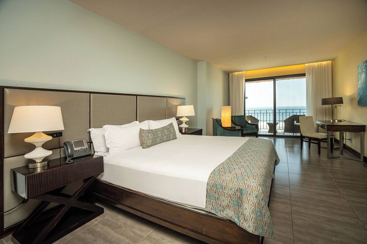 Ein Hotelzimmer mit einem großen Bett, Nachttischlampen, einem Schreibtisch mit Stuhl und Meerblick durch das Fenster.