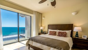 A spacious bedroom with a large bed, two bedside tables with lamps, a view of the ocean through floor-to-ceiling windows, and a ceiling fan. - Crocs Resort & Casino (Jacó)