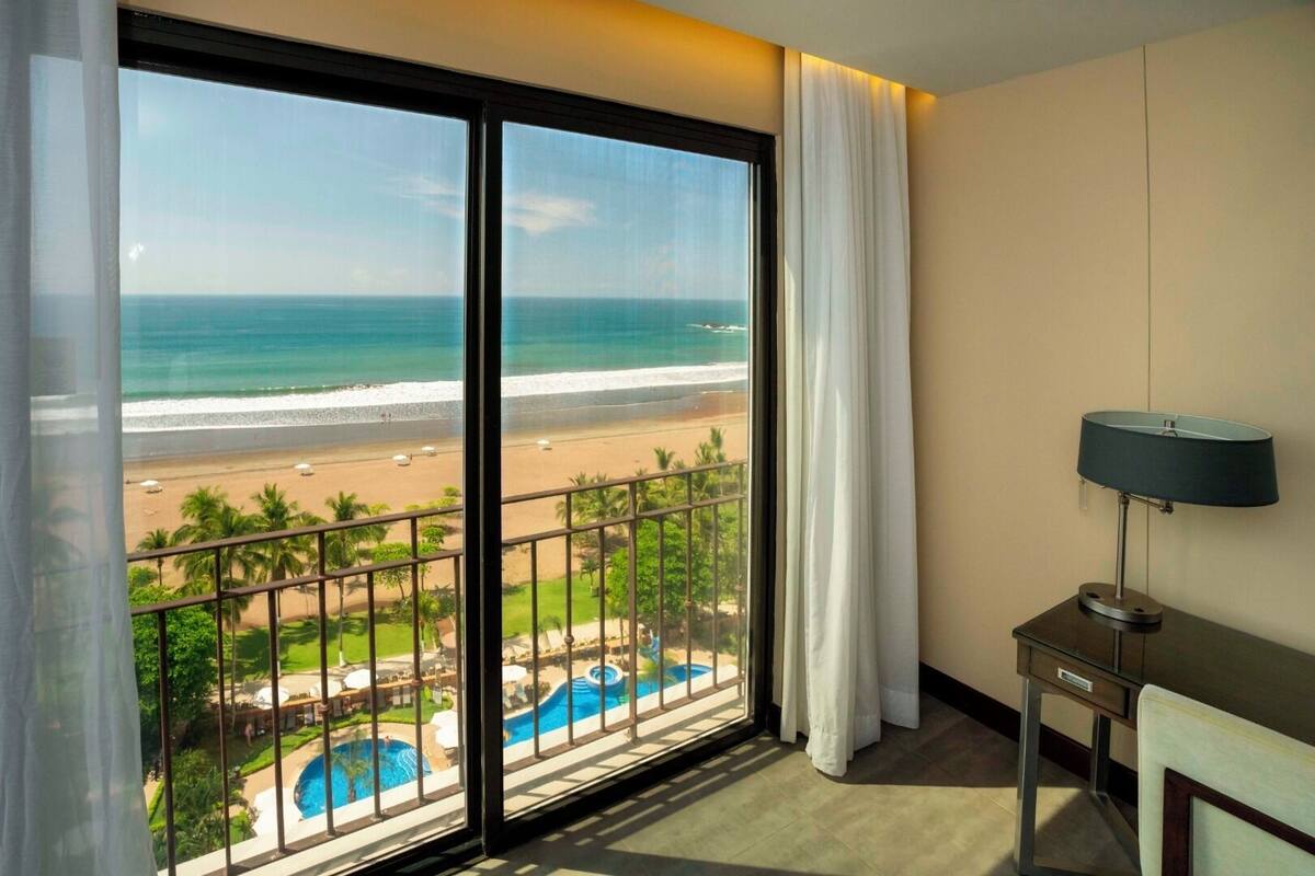 Ein Hotelzimmer mit einer großen Schiebetür, die auf einen Balkon mit Blick auf Strand und Pool führt.