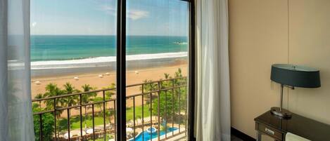 Habitación de hotel con un amplio ventanal corredero que da a un balcón con vistas a la playa y la piscina.