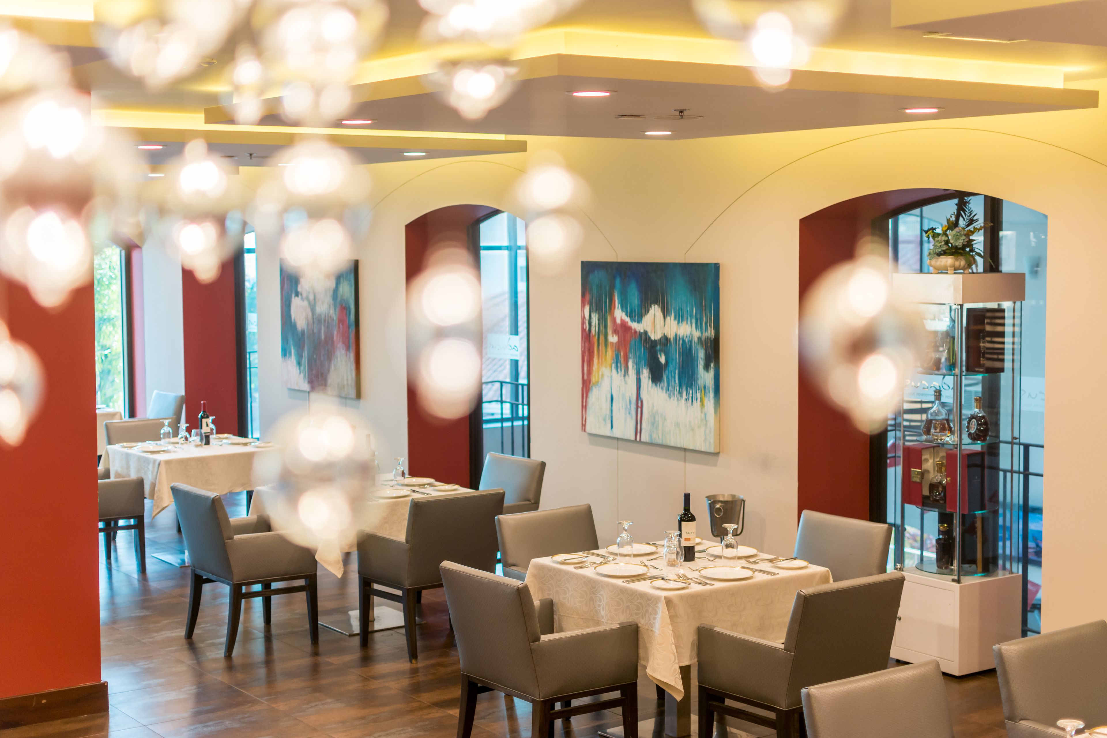 A modern dining area with tables set for four, large windows, and a glass display case with bottles.