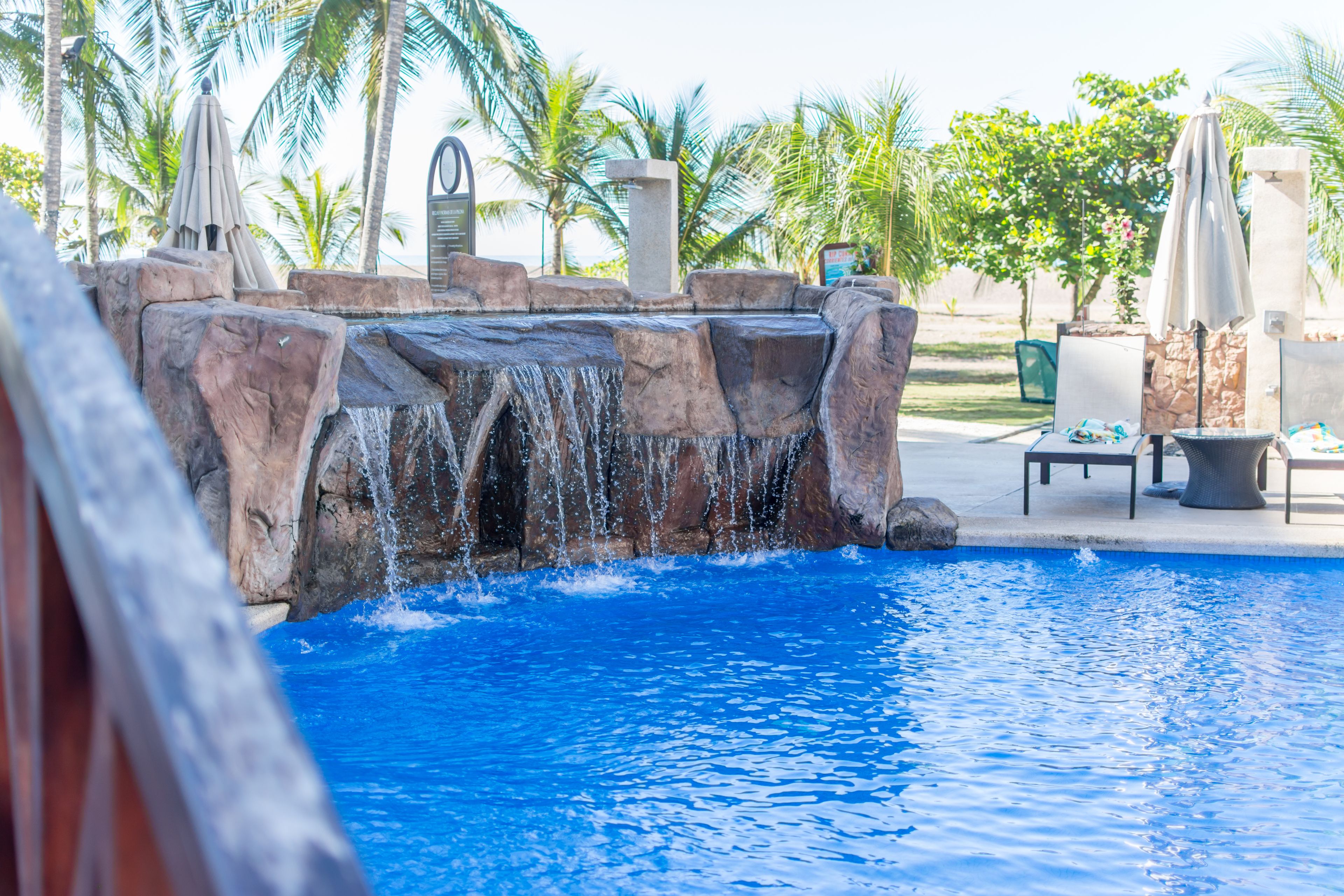 A swimming pool with a waterfall feature, surrounded by palm trees and outdoor seating.