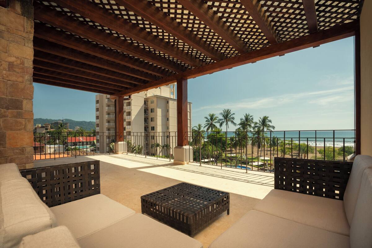 Ein Balkon mit Blick auf den Strand und Palmen, ausgestattet mit einer Couch und einem Couchtisch.