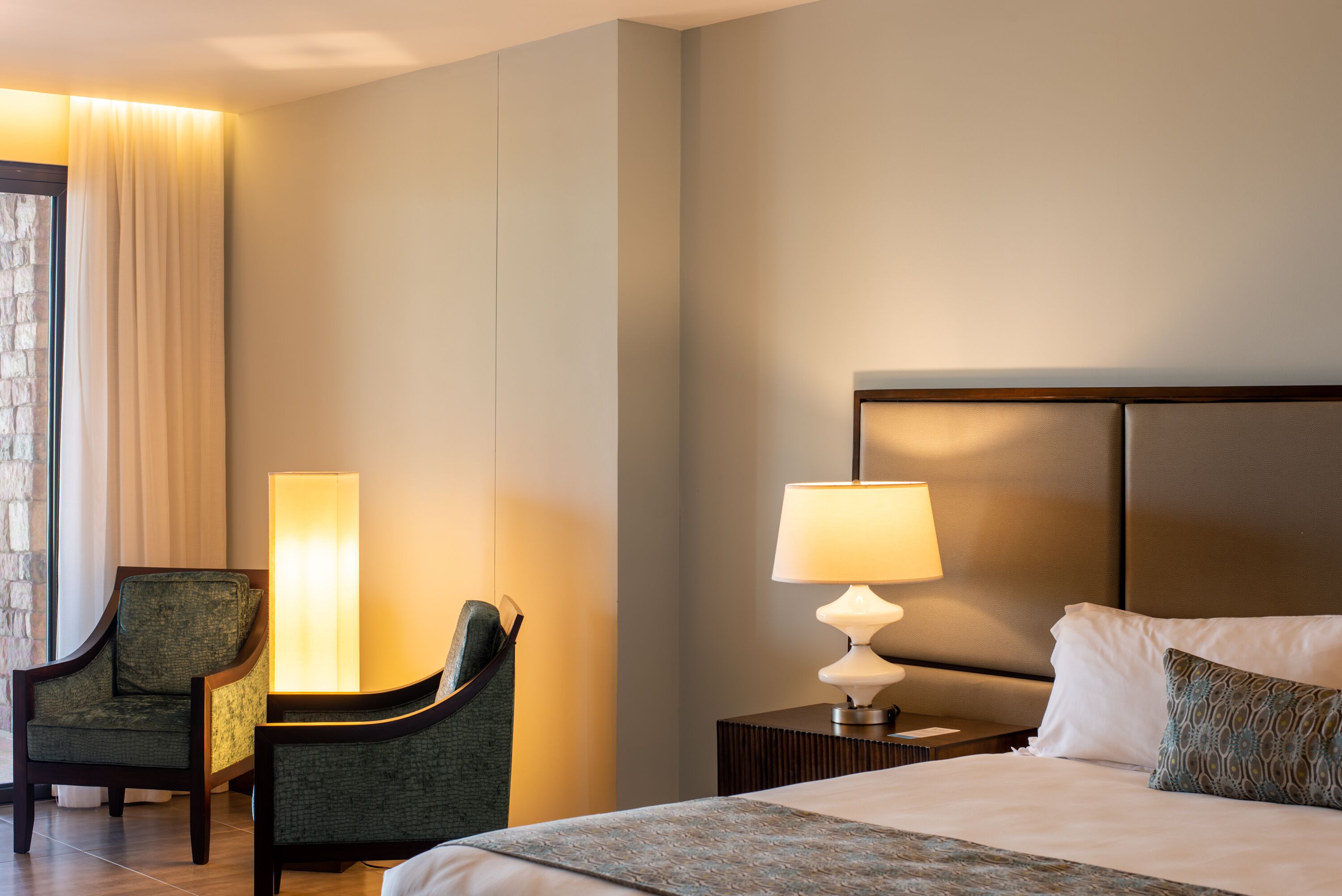 A hotel room with a bed, a nightstand with a lamp, a chair, and a large window with curtains.