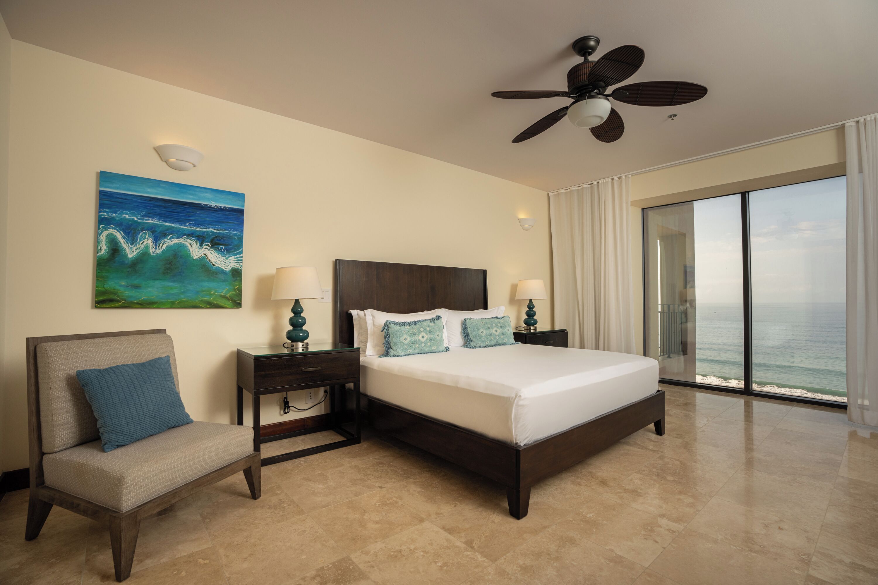 A hotel room with a large bed, a chair, a painting of the ocean, a ceiling fan, and a view of the ocean through large windows.