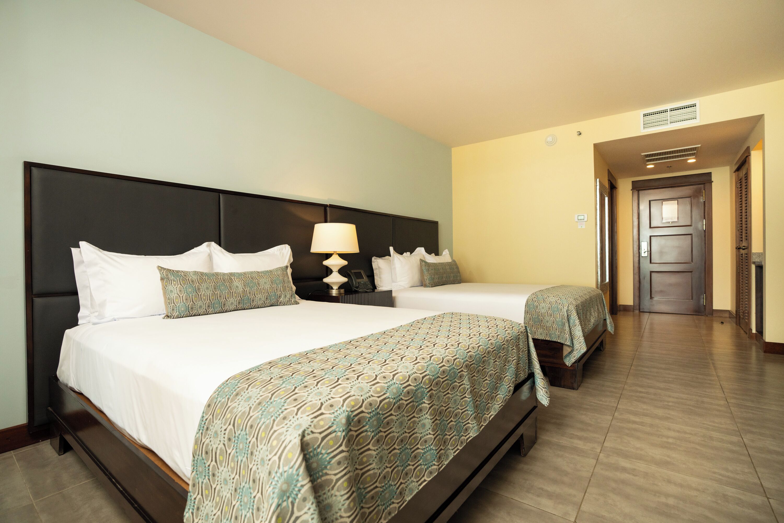 A hotel room with two beds, a nightstand with a lamp, and a door leading to another room.