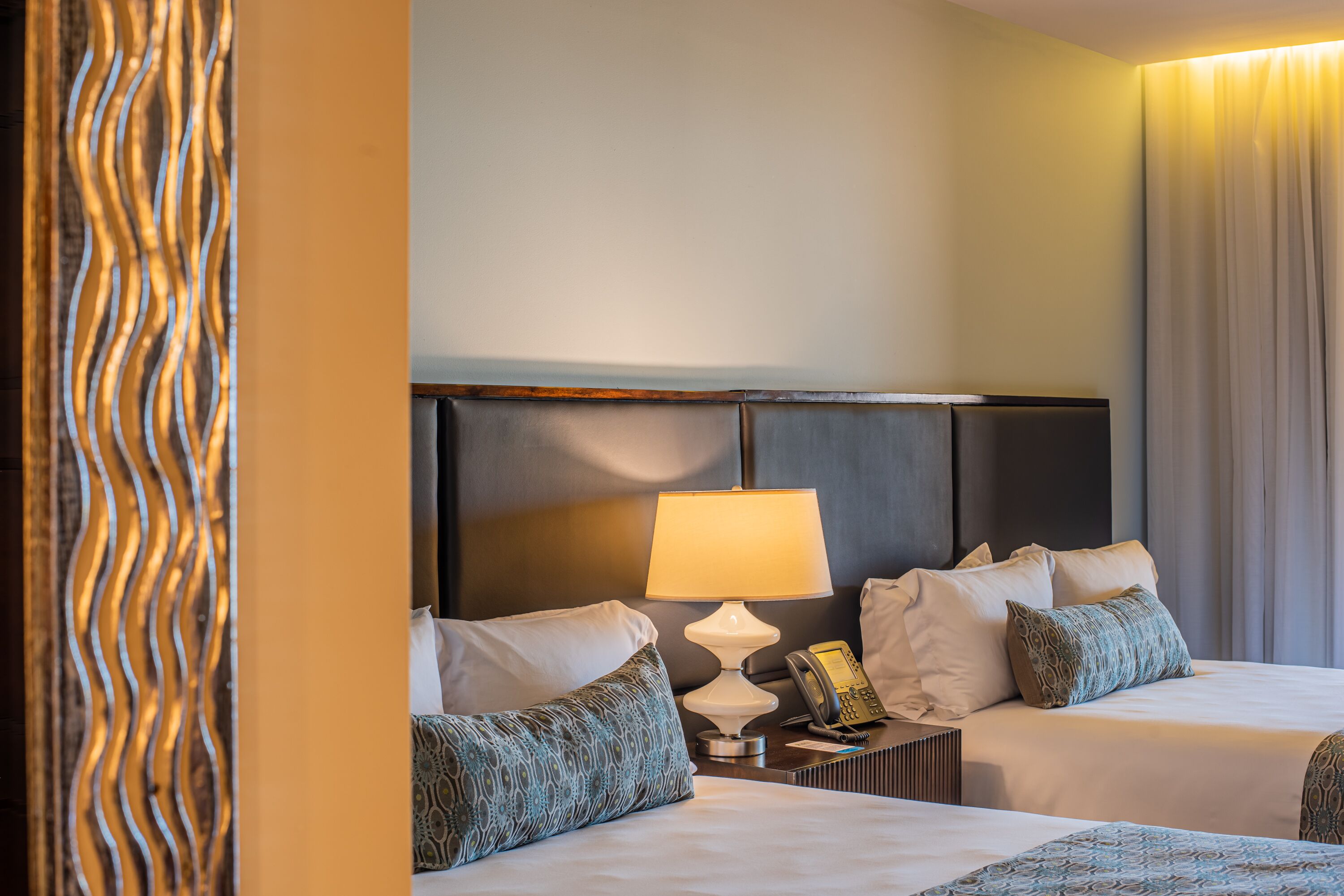 A hotel room with two beds, a bedside lamp, a telephone, and a decorative wall panel.