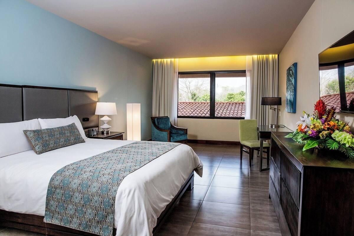Ein Hotelzimmer mit einem großen Bett, einem Schreibtisch mit Blumen, einem Sessel und Blick ins Freie.