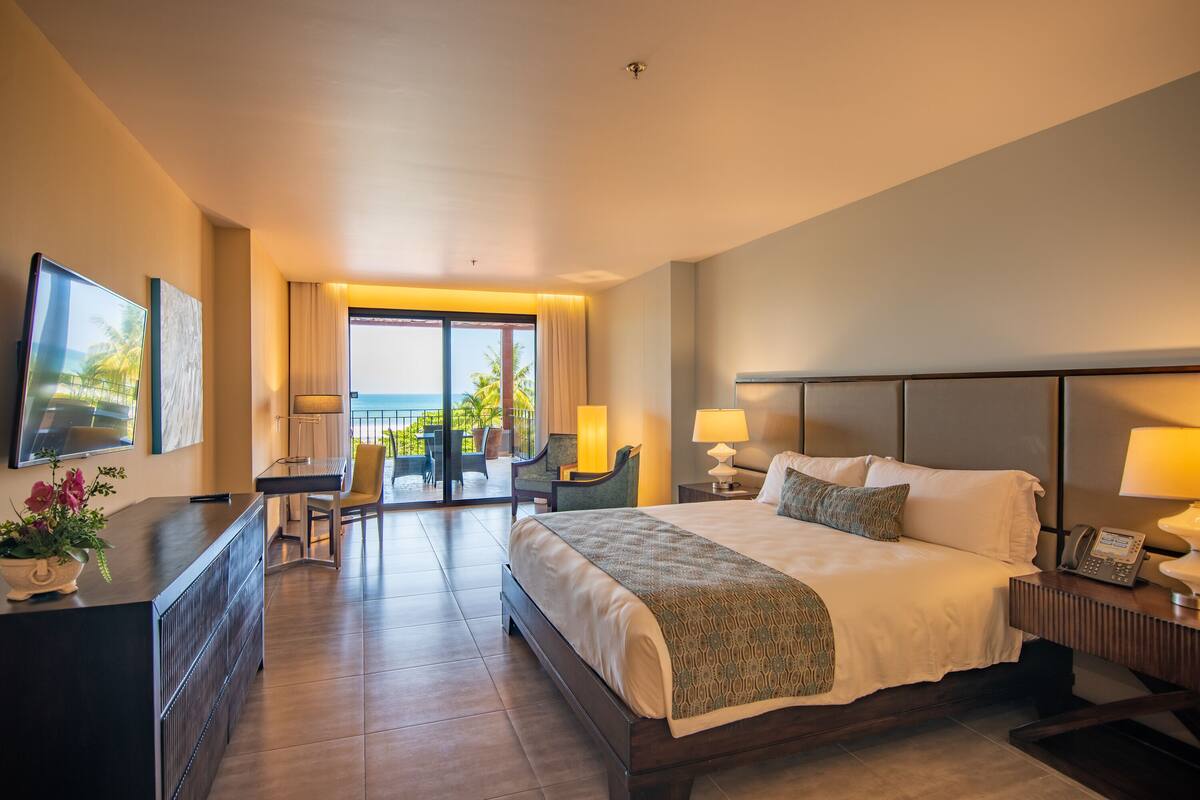 Ein Hotelzimmer mit einem großen Bett, einem Schreibtisch mit Stuhl, einem Fernseher und einem Balkon mit Meerblick.