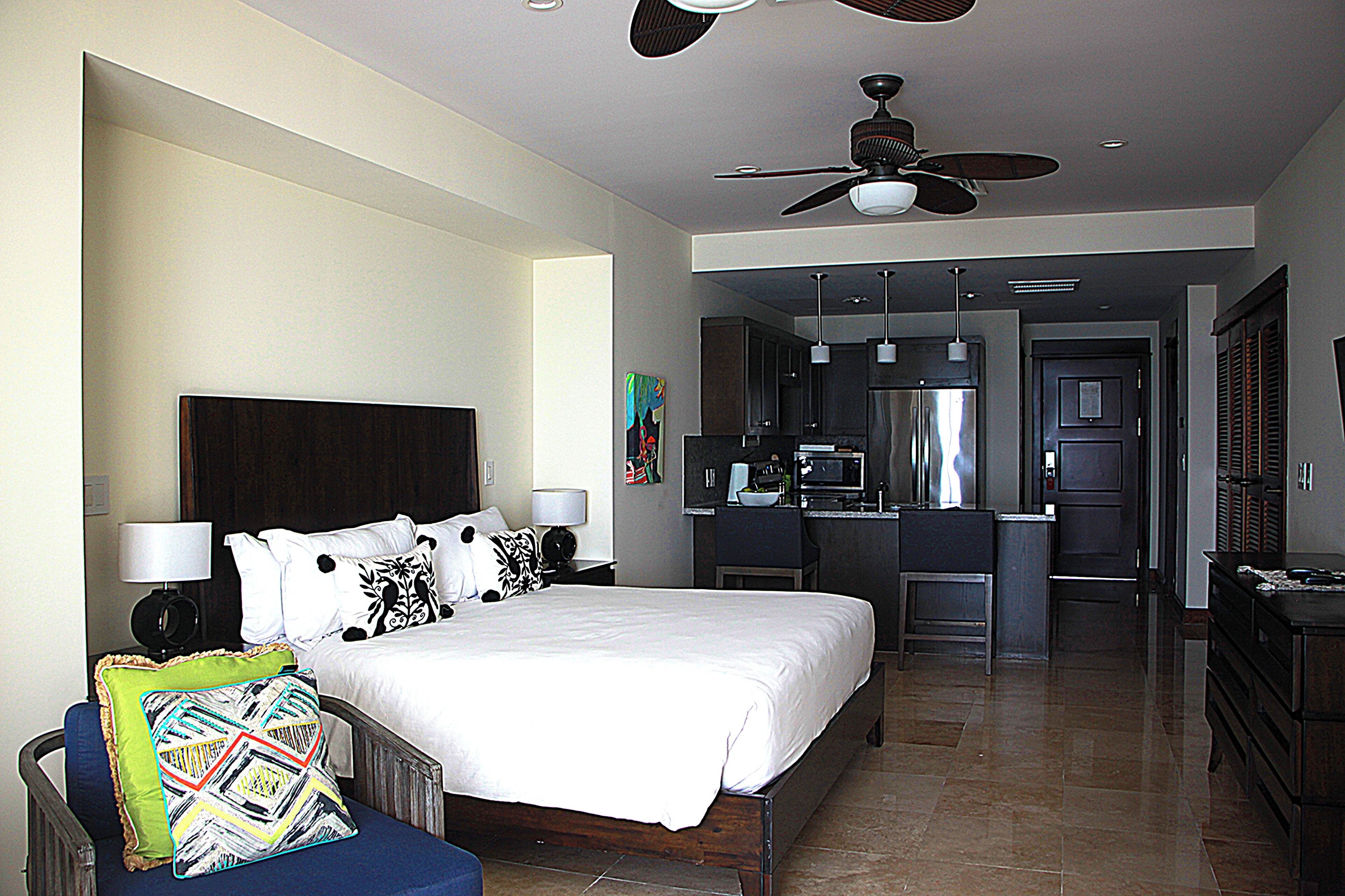 A hotel room with a large bed, a desk, a kitchenette, and a ceiling fan.