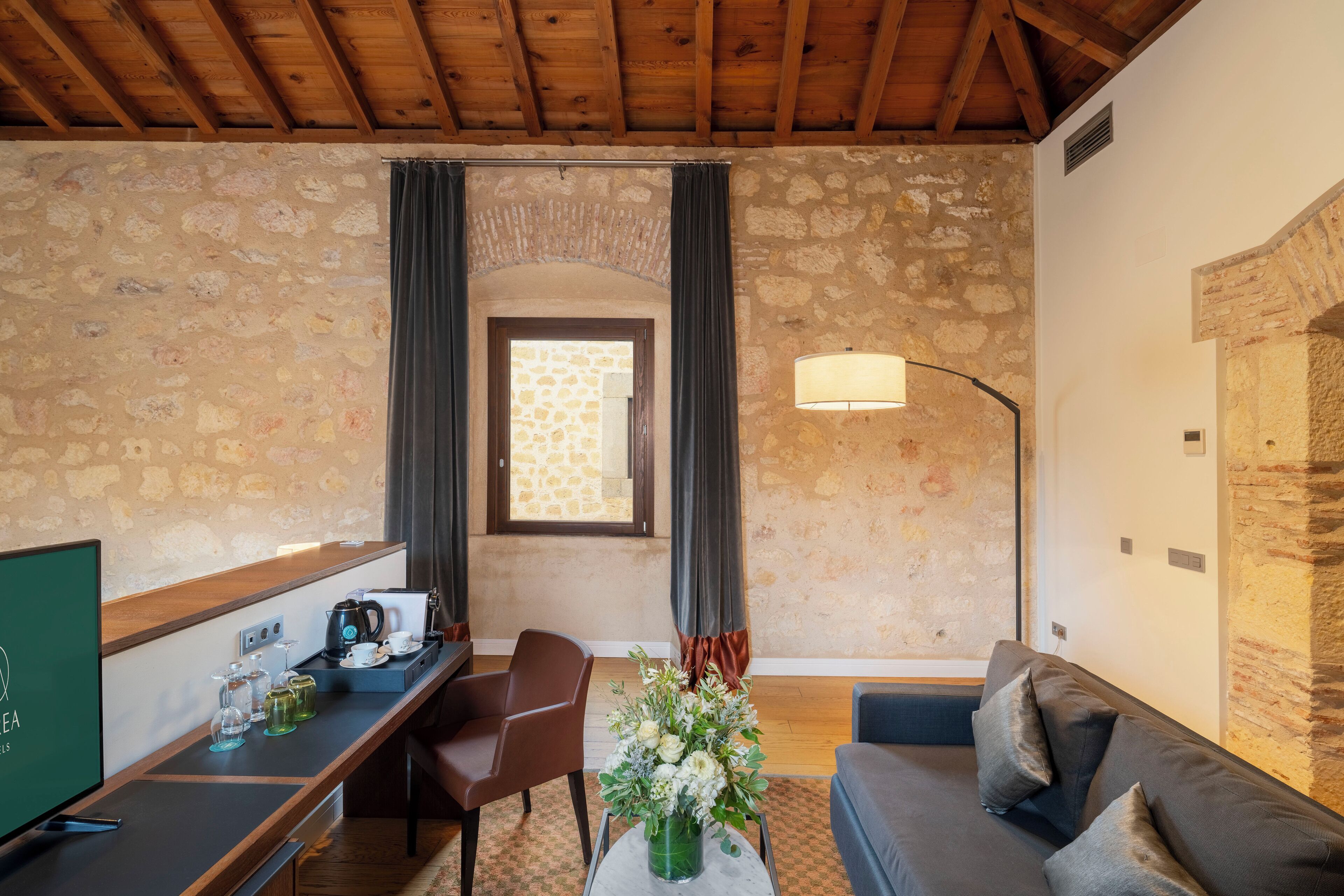 Una habitación acogedora con paredes de piedra, techo de madera, un escritorio con cafetera, un sofá y una lámpara.