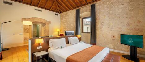 Una habitación de hotel con techo de madera, paredes de piedra, una cama grande con una manta marrón, un televisor y una mesita de noche con una lámpara.