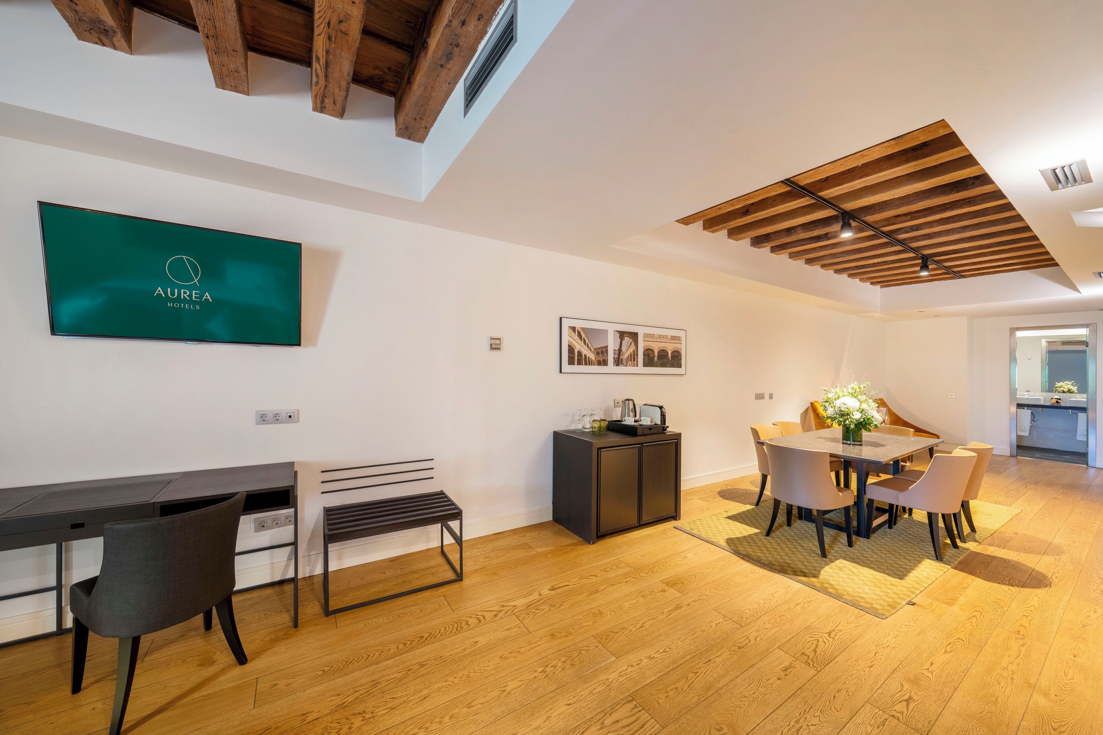 A modern hotel room with a wooden ceiling, a dining area with a table and chairs, a bench, a cabinet, and a TV mounted on the wall.