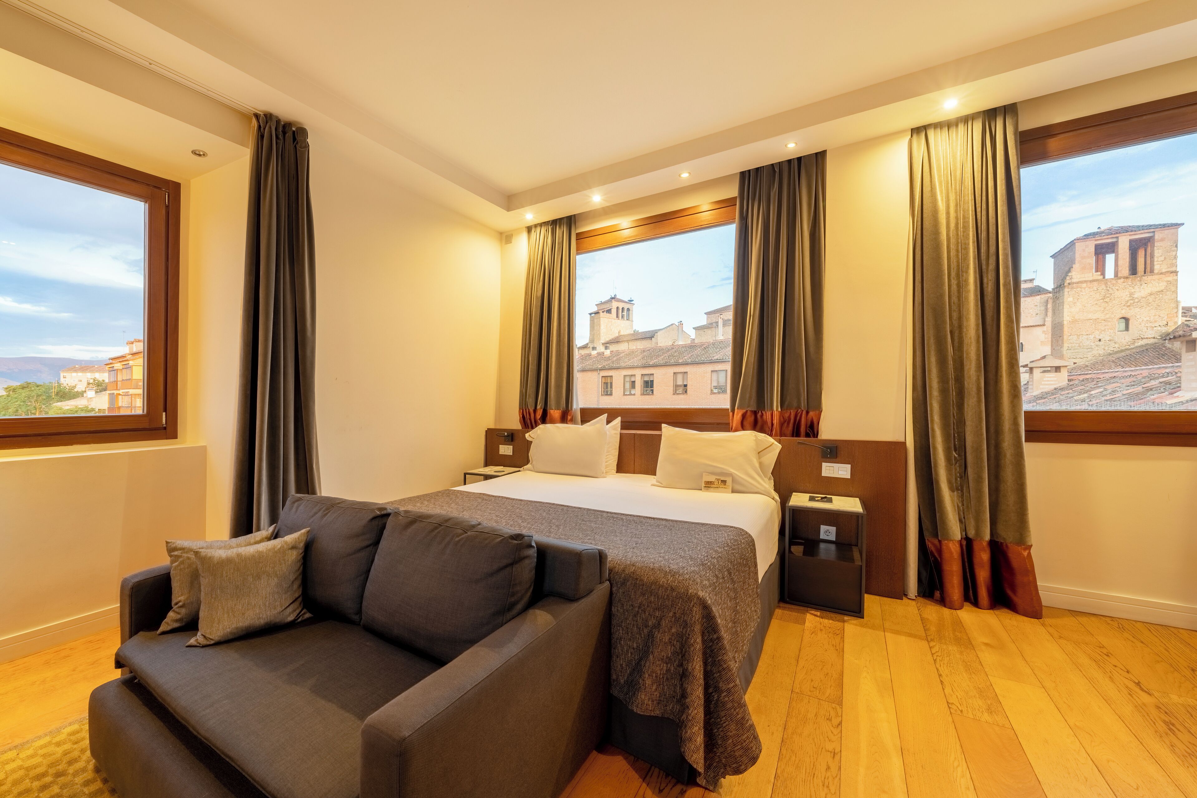 Quarto de hotel com cama, sofá e amplas janelas com vista para a cidade.