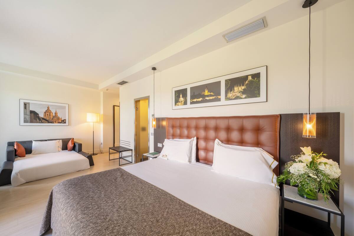 Een hotelkamer met een groot bed, een kleiner bed, een nachtkastje met een bloevas, een lamp en ingelijste foto’s aan de muur.