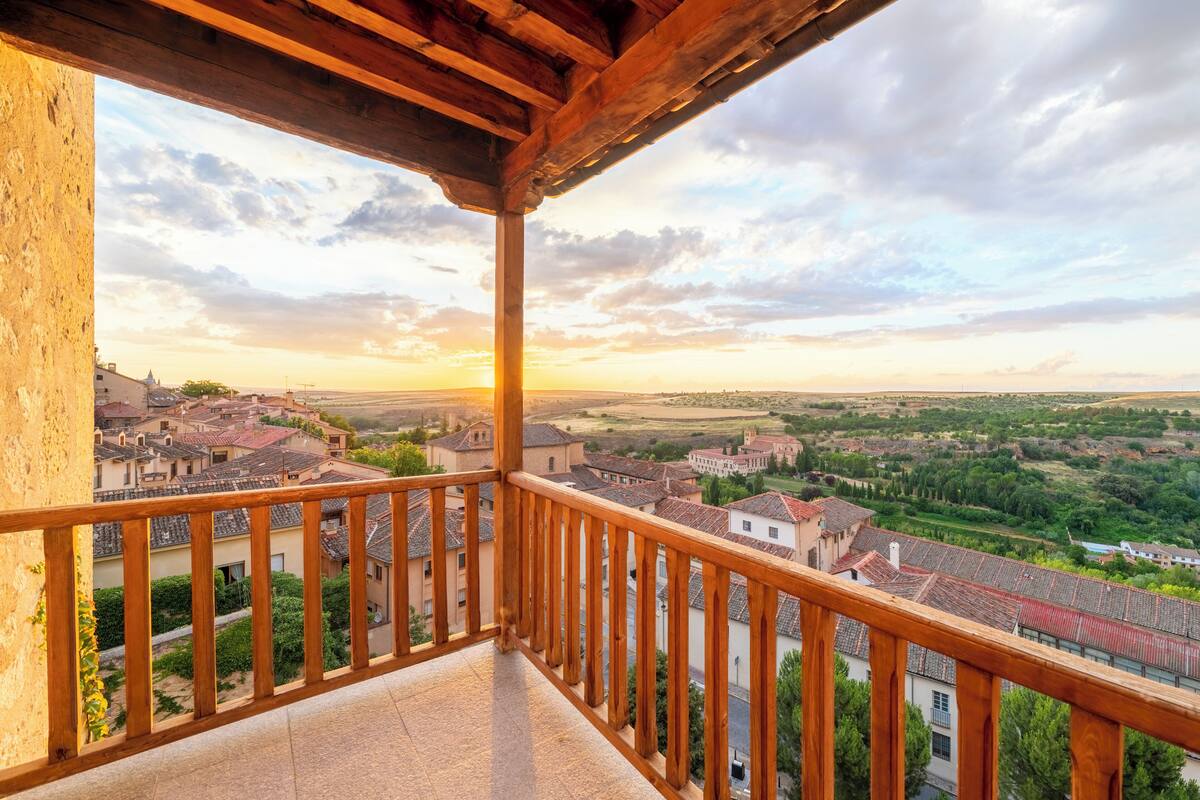 Een balkon met houten hekwerk met uitzicht op een stad met rode daken en groene velden.