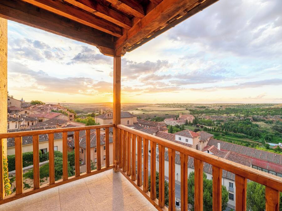 A balcony with a wooden railing overlooking a town with red-roofed buildings and green fields.