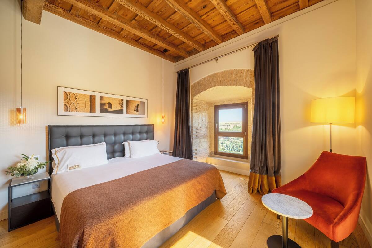 Een hotelkamer met een groot bed, een rode stoel, een klein tafeltje en uitzicht op een stenen boog.