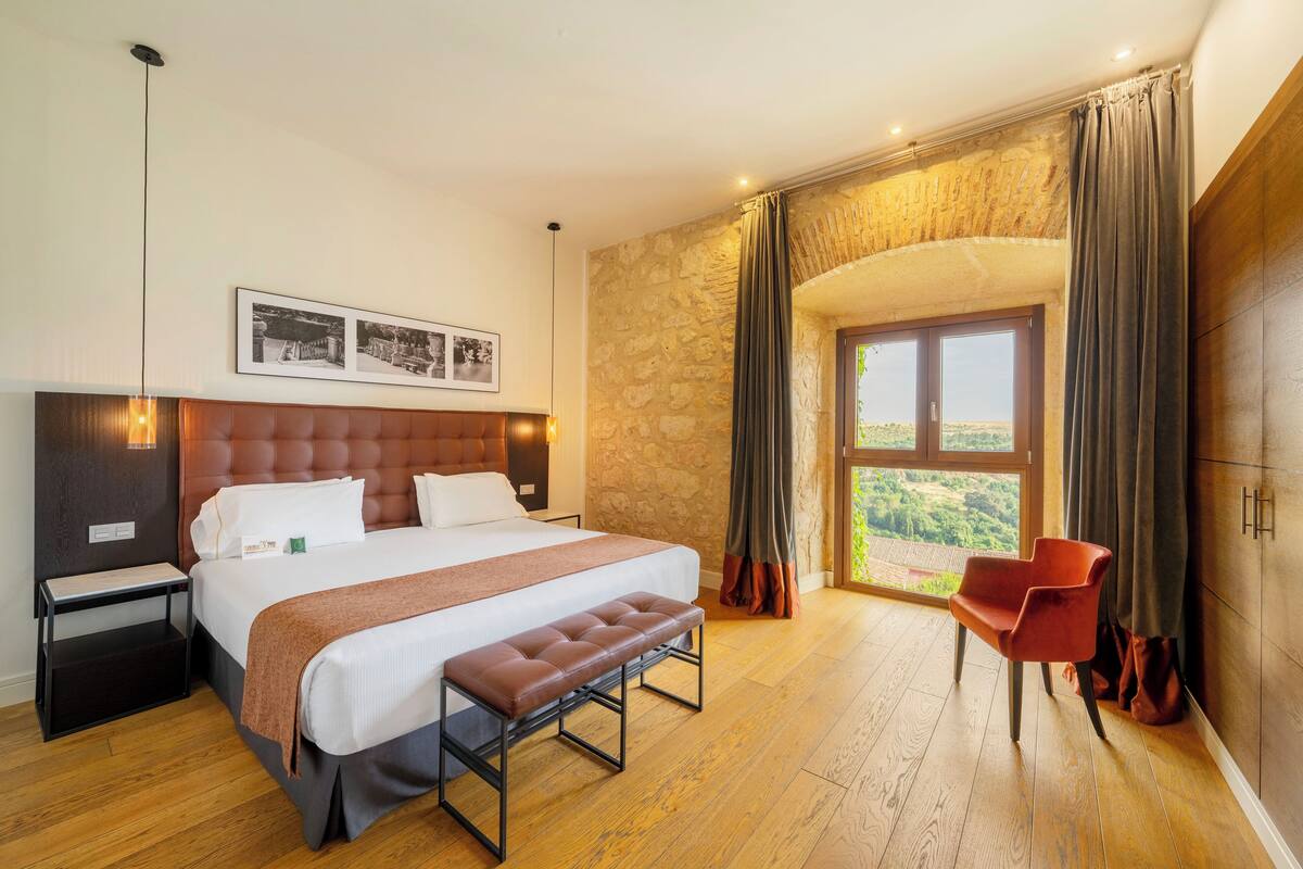 Een slaapkamer met een groot bed, een bankje, een nachtkastje, een stoel en een stenen muur.