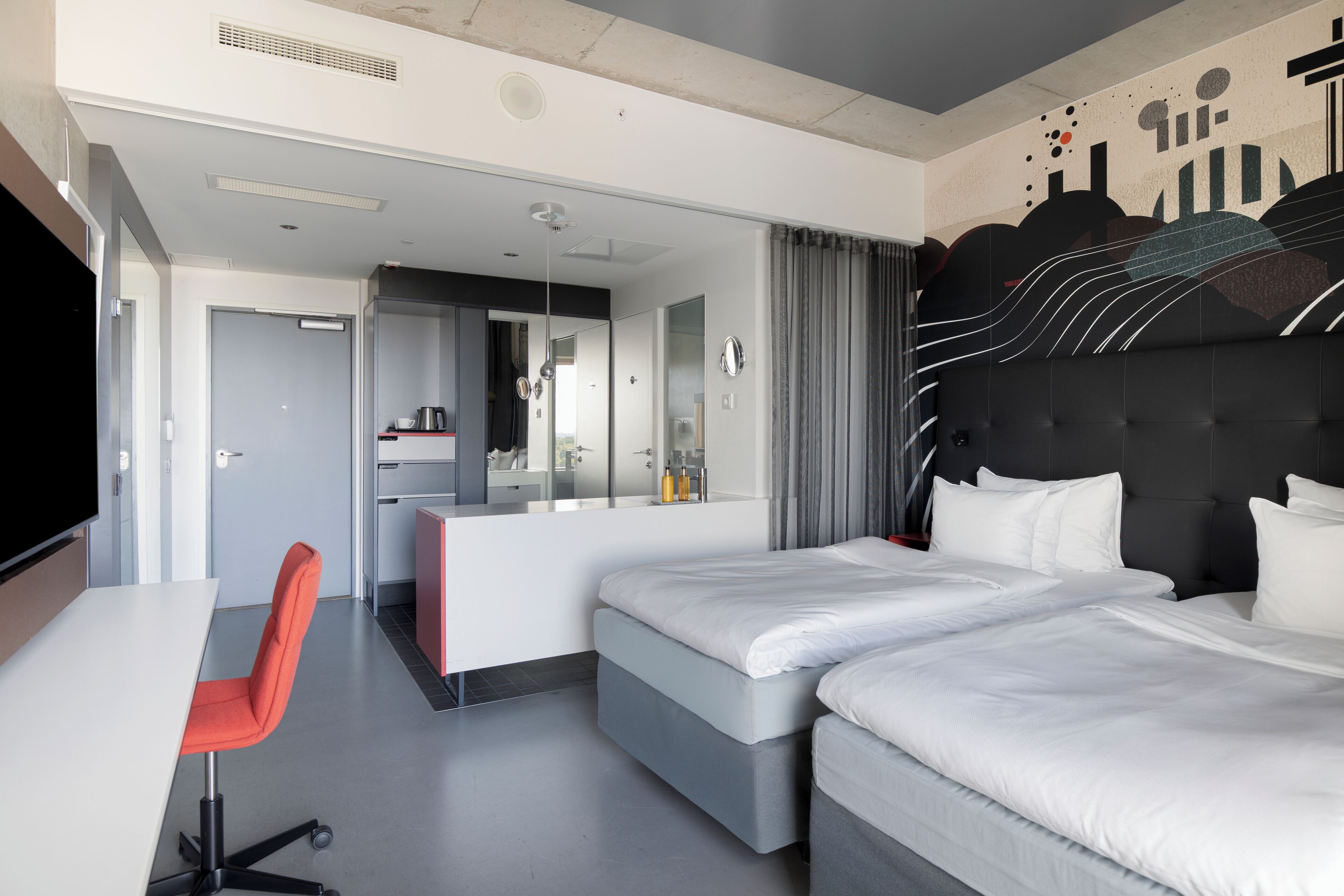 A modern hotel room with a large bed, a desk, and a chair. There is a bathroom with a mirror and a shower.
