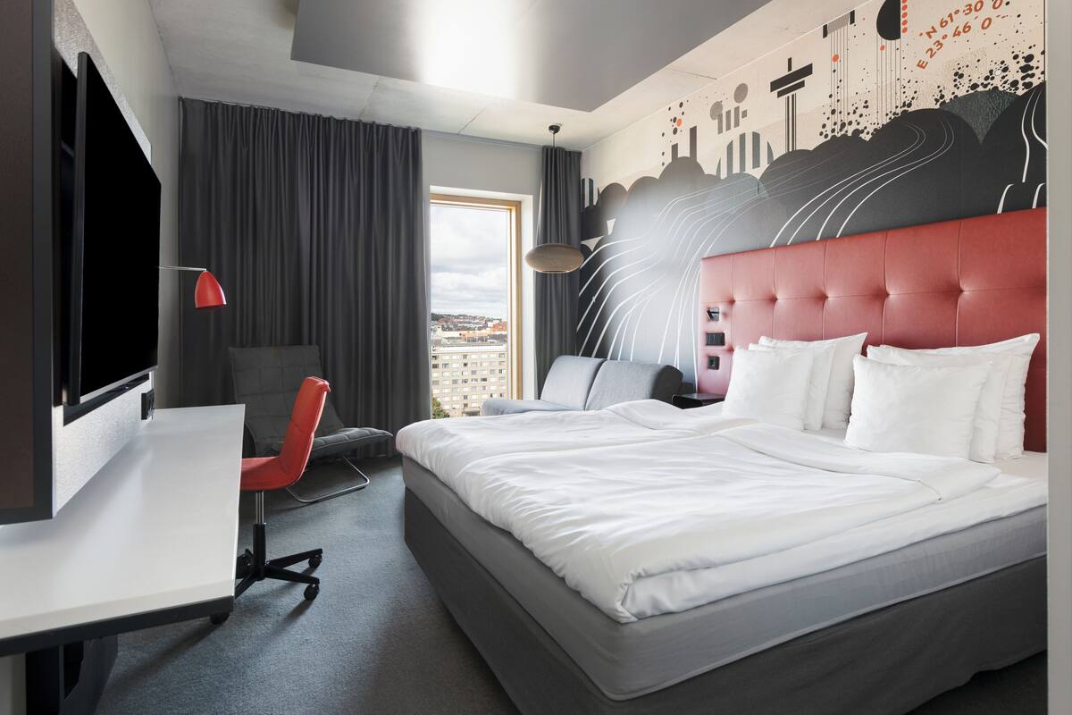 A modern hotel room with a large bed, a desk, a chair, and a city view through the window.