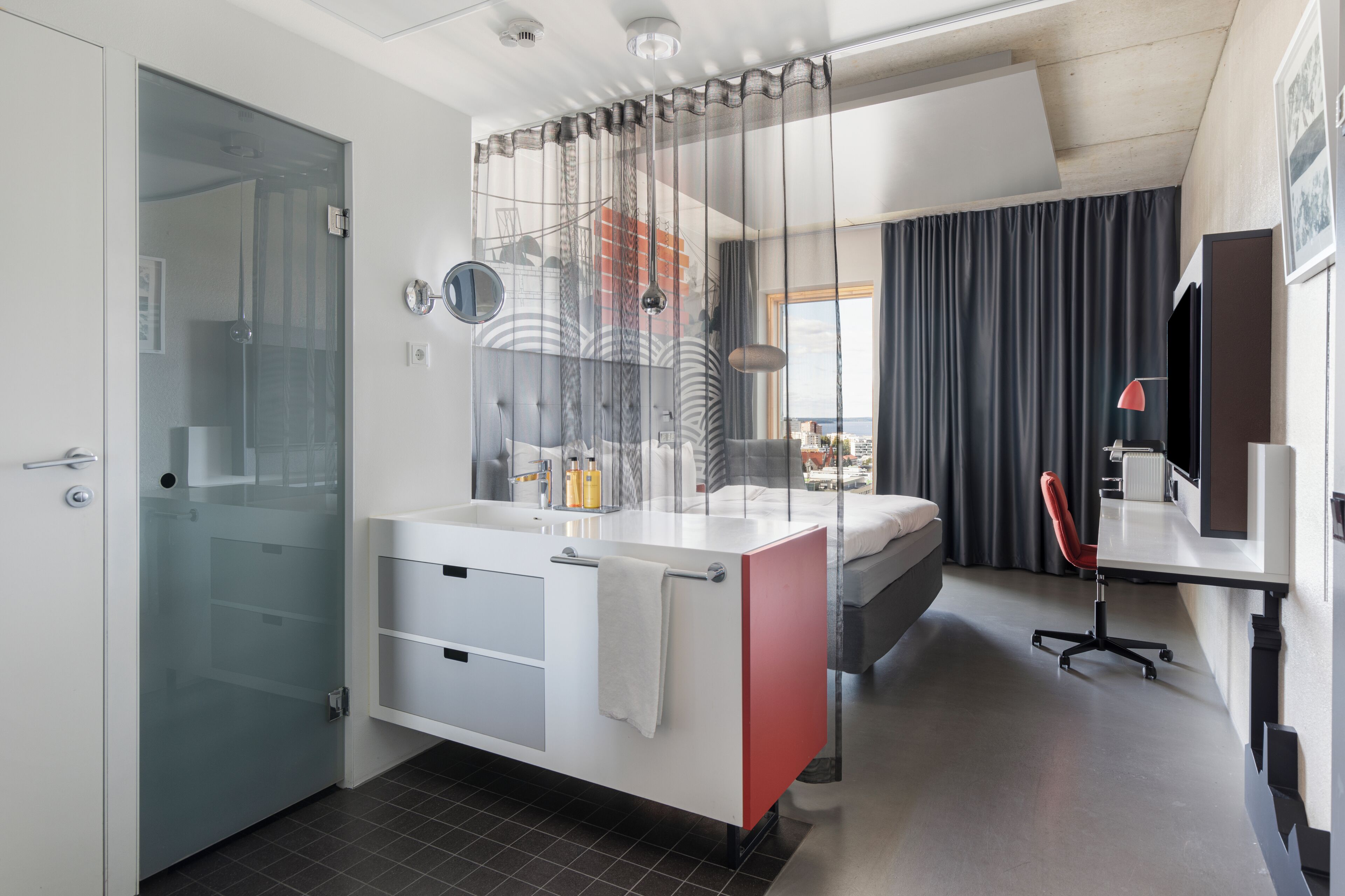 Un baño moderno con cabina de ducha de vidrio, un lavabo blanco con detalles rojos y una cama con vistas a la ciudad.